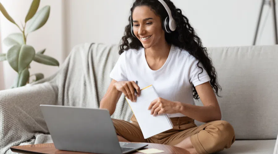 A young girl smiling and sitting on her couch with headphones on and on her laptop. She is in an online course for school and taking notes in her notebook.