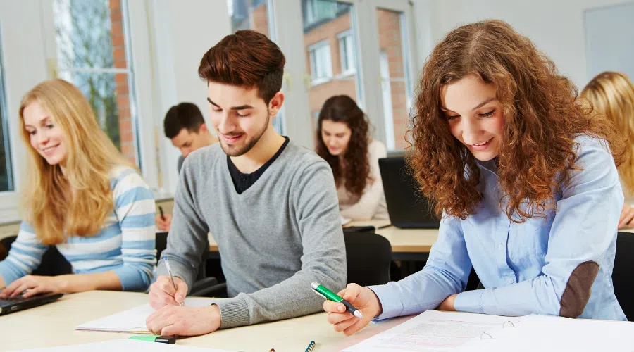 Three college students at a table taking notes with a pen in their notebooks. They are enrolled in a course for college or high school.