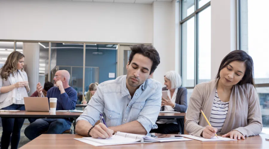 Two adults in the forefront taking hand written notes during a course. In the back an older man is arguing with a women over his computer.