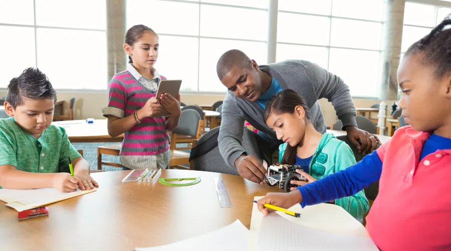 A group of kids working on a robotics program, writing notes on paper, and talking with a teacher about a project.
