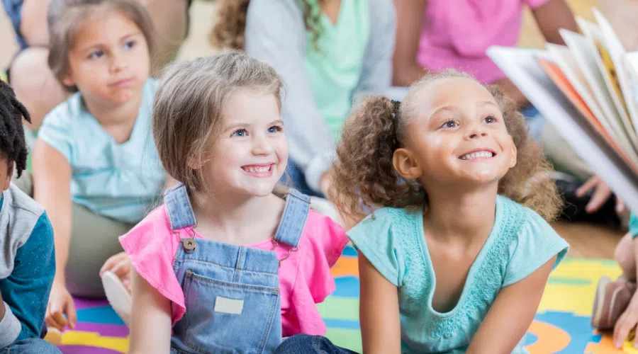 two girls sitting in a classroom setting smiling while someone reads a book in the foreground