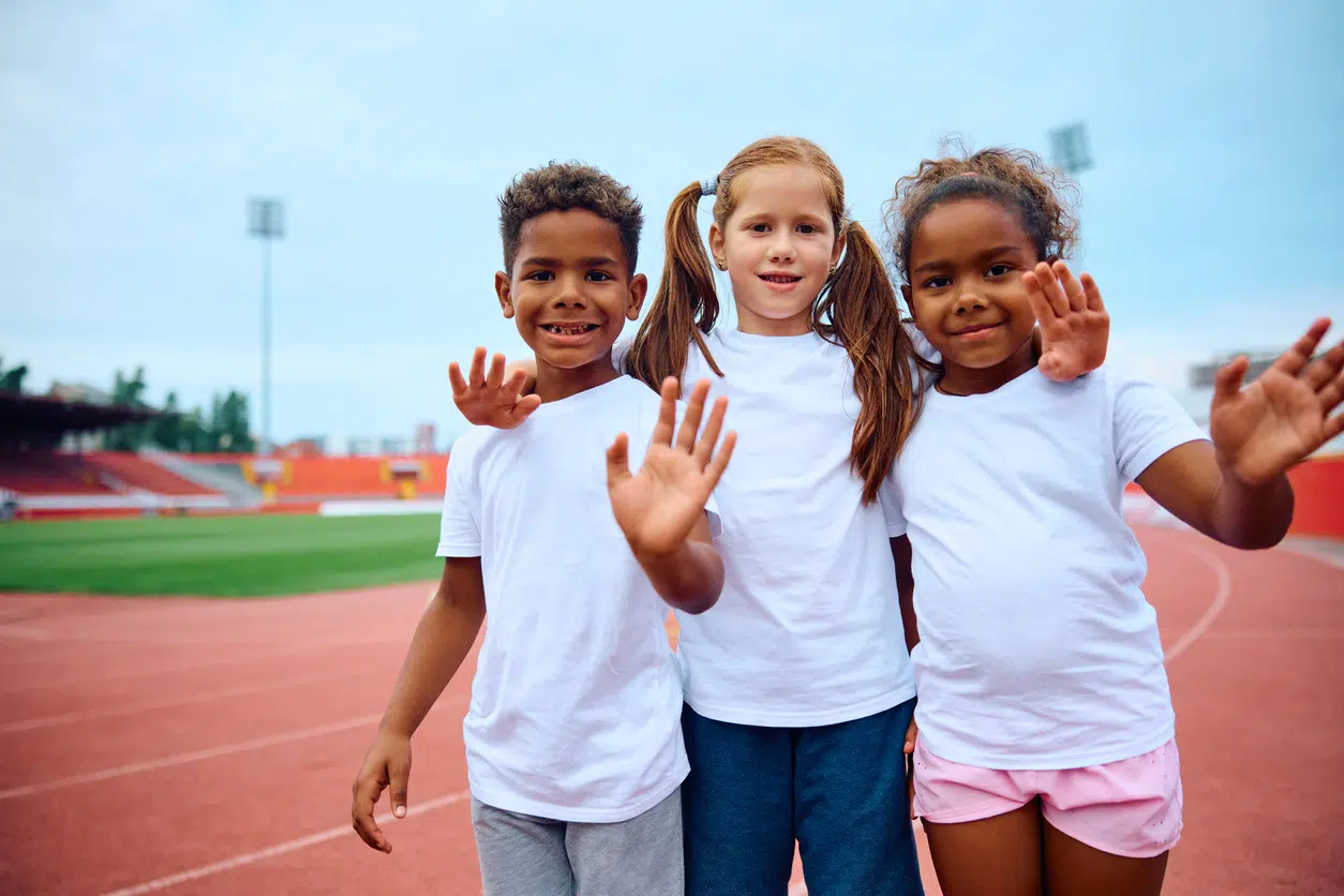 Happy little friends embracing waving during PE class at the stadium and looking at camera.