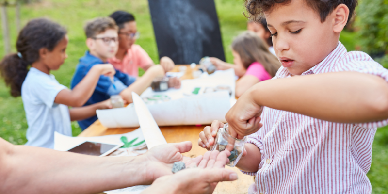 kids sitting around a picnic table doing an art project with one kid handing an adult rocks from a jar