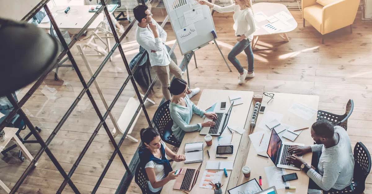 an overhead shot of a modern office, three people working at a computer, two standing displaying a graph