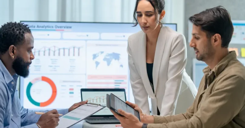a woman and two men looking at reports on tablets with a large screen of graphs behind them