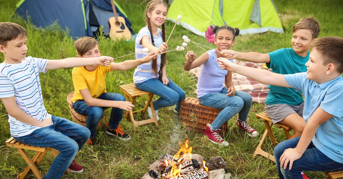 Six kids roasting a marshmallows surrounded by camping tents in the grass