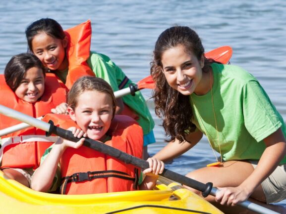 Three young kids in orange life vests sitting in a yellow kayak with a camp counselor on the side steadying the boat