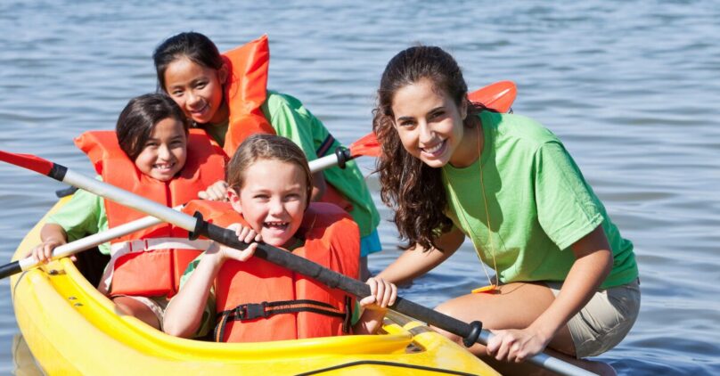 Three young kids in orange life vests sitting in a yellow kayak with a camp counselor on the side steadying the boat
