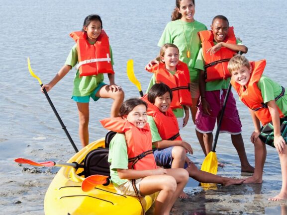 five kids with orange life vets on in a lake, sitting on a yellow kayak with an adult hugging two kids